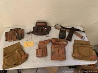 Full view of the seven vintage leather tool belts and pouches arranged on a table, showing variety in size, color and pocket design.