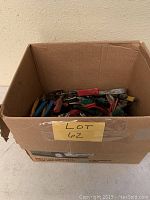 Full cardboard box filled with assorted pliers and hand tools with plastic handles in various colors, showing quantity and condition.