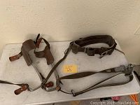 Pair of pole climbing spikes with leather padding, metal frame, and leather straps laid on a table alongside two leather tool belts with metal buckles.