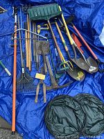 Top-level view of various garden tools including shovels, rakes, hoes, and pruning shears laid out on a blue tarp with two black collapsible containers visible.
