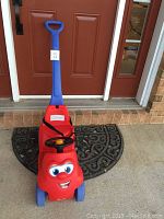 Front view of the red ride-on toddler toy car with smiling face and blue removable push handle attached at the back.