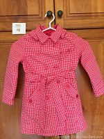 Full front view of the girls rain coat hanging on a white hanger showing its red and white checkered pattern, double-breasted button front, collar, and belt.