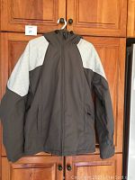 Full front view of grey Billabong ski/snowboarding jacket with lighter panel accents on shoulders and hood, hanging on wooden cupboard. Shows zippered front, pockets, and overall style.