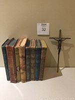 Side view of eight vintage hardcover books with worn edges and an INRI cross positioned to the right on a flat surface against a wall.