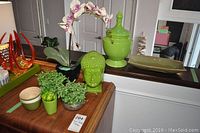 View of four faux plants including a faux orchid, green Buddha head sculpture, green lidded vase, and green rectangular plate.