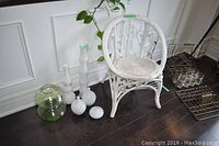 Photo showing white wicker chair and six glass vases in front of a white paneled wall on dark wood floor