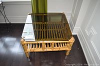 Top, angled view of square bamboo frame coffee table with dark tinted glass top and bamboo lower shelf on dark hardwood floor against white wall and green curtain