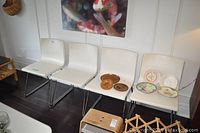 Four white chairs with faux leather seats and backs on metal sled frame bases. They are lined up against a wall with a painting above. The chairs appear lightly used with some wear visible.