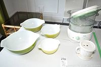 Photo showing four vintage Pyrex Cinderella bowls in Green Verde pattern alongside a T Fal steam cuisine appliance and a small Corning Ware mug with lid.