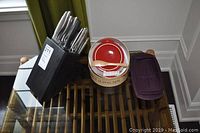KitchenAid knife set in black wooden block with sharpener, Brie Baker ceramic dish with red interior and wooden spatula, and Pampered Chef silicone steamer with lid on a glass table.