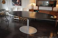 Full view of modern oval pedestal table showing dark wooden top with smooth finish and silver metal base in room setting.