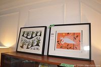 Two framed art pieces resting on top of a cabinet under a wall molding; one features an abstract tree design, the other a white bird on orange fabric.