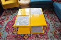 Four yellow metal square side tables arranged together on a colorful patterned rug. Two have marble inserts on top, two have solid yellow metal tops.