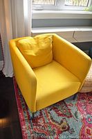 Yellow fabric armchair with cushion, positioned on a colorful rug near window and side table.