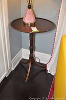 Round wooden side table shown with pink lamp base, tripod leg base visible and signs of wear on the wood surface.