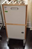 Front view of the filing cabinet showing the top hanging file drawer with metal handle and bottom lockable cabinet door in white with natural wood frame and legs.