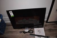 Wall hanging black electric fireplace insert with glass front, power cord and mounting bracket on wood floor.