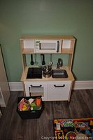 Front view of wooden Ikea toy kitchen set showing microwave, stovetop, sink, cabinet and drawer with metal pots and pans on countertop and utensils hanging above