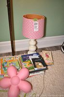 Full view of pink table lamp with white base and geometric shade, children’s books stacked beside it on carpet
