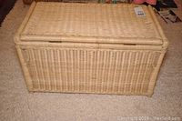 Front view of light beige wicker storage trunk on carpeted floor showing rectangular shape and woven texture.