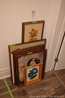 Stack of five framed embroidered and crewel wall art pieces leaning against a wall showing different sizes and wood frame colors.
