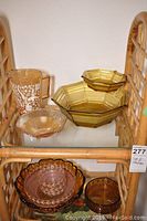Photo showing two-tier amber chip and dip bowls, footed glass bowl, pitcher with floral pattern, and small glass bowls on a rattan shelf.