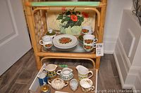Photo showing the entire assortment displayed on a wooden shelving unit, including teacups, plates, creamers, sugar bowls, and the poppy painting in the background