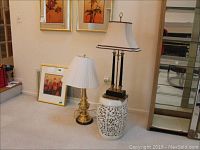 Two brass table lamps side by side on carpet, one with marble base, one with carved white ceramic base, both with fabric shades