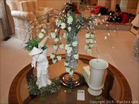 Full view of the three main items on a round table in a living room: ceramic Asian man figurine, glass vase with faux flowers, and white Lenox crackle ceramic vase