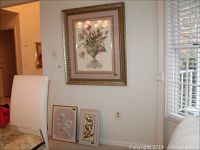 Overview of three framed wall art pieces as displayed on and near a wall beside a window. Large floral print hangs on wall, smaller two pieces lean against the wall on the floor.