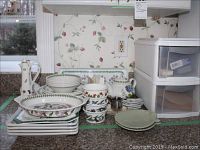 Photo showing stacked Botanic Garden dinner plates, cereal bowls, custard cups, teapot, vinegar and oil set, ceramic teabag holders, and additional plates on a kitchen counter with floral wallpaper.