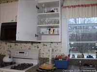 Kitchen cabinet with glassware including juice glasses and mugs on shelves, white microwave, stove, white kettle, paper towel holder, and baskets with kitchen linens.