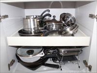 Full view of a cabinet drawer with nesting cookware including stockpots, saucepans, fry pans with black handles, metal lids and glass lids; electric broiler seen at bottom right.