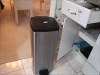 Photo of Brabantia stainless steel step-on kitchen trash bin with black lid and foot pedal, placed on kitchen floor near cabinet