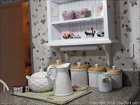 Shelf with four white ceramic canisters with wooden lids, one white ceramic pitcher, jar candle, small ceramic statue, covered tureen with ladle and wind-up flashlight on table