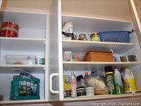 Kitchen cabinet shelves with bottles of cleaning supplies, spray cans, plastic containers, detergent bottles, cleaning cloths, and small baskets.