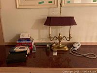 Desk setup with the brass bankers lamp, vintage stapler, tape dispenser, stack of books, and corded telephone displayed on a wooden surface.