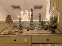 Full view of dresser with all items including lamp, obelisks, small figurines, cross plaque, and large covered vase with faux flower arrangement on top
