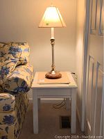 Lamp with brass and wood base and white shade on white wooden table next to floral couch