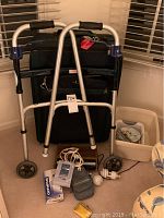 Photo shows silver aluminum Drive walker with black foam handles and wheels in front of a black Atlantic suitcase with wheels. Various smaller items including camera accessories and storage bins are at the base.