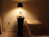 Brass candlestick lamp on black pedestal with white cushioned vanity bench nearby, lamp lit