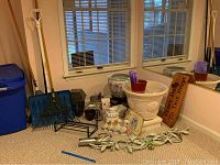 Photo of various garden tools and decorative items against window with blinds. Included are rakes, a snow shovel, mosquito zapper, large ceramic pots, porcelain pot on stand, small ceramic pots, and decorative wooden signs.