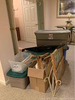 Photo shows a group of three large plastic storage bins with lids in gray and green colors, a cardboard box with clothes hangers (wooden and plastic), and a collapsible wooden and mesh pet gate placed on carpeted floor.