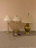 Frontal photo of three brass tone lamps with pleated white shades showing two taller slender lamps and one shorter lamp with wider base, cords visible on carpeted floor, beige wall background.