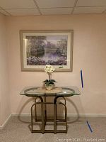 Photo showing brass and glass accent table with vase and faux white flower plant on top, with framed landscape print mounted on wall behind.