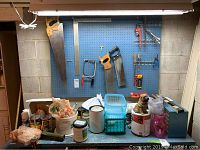 Pegboard with saws, clamps, wrenches including two hand saws, hacksaw, pipe wrench and pliers. Shelf below holds paint cans, paint tools, storage containers, and various hardware items.
