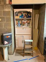 Wooden white cabinet with cleaning supplies on shelves, pull down cabinet section, and lower storage doors. Also visible small table with paper shredder and wooden step ladder beside the cabinet.