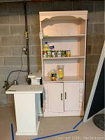Tall white wooden cabinet with two shelves, two-door lower storage cabinet, holding canned goods and pantry items, photographed in front of cinder block wall.
