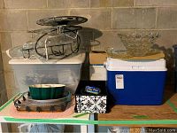 Overall lot showing Rubbermaid cooler, glass bowls, Bundt pan, casserole dish, decorative plate, and oven bags on table in basement.