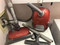Overall view of the Electrolux red canister vacuum and attached hose, placed on laundry appliances with cleaning attachments spread around.